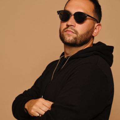 Man with short dark hair wearing black sunglasses stands with arms folded